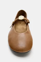 Zapato tipo bailarina de piel color marrón tostado, con tira delantera ajustable, aplique metálico y punta redonda.