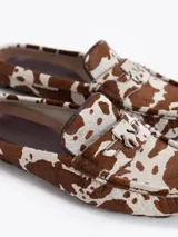 Mocasín destalonado con estampado animal print de vaca en tonos marrón y blanco, confeccionado en pelo vacuno. Presenta un aplique metálico en la capellada y suela de caucho.