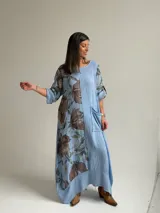 Vestido largo celeste con gran estampado floral marrón en el lateral derecho, de corte holgado, mangas 3/4 con trabilla y dobladillo irregular con flecos desflecados. Incluye un bolsillo aplicado en el frente.