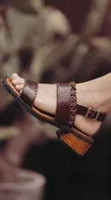 Sandalia de cuero color marrón, con taco bajo de madera, tiras anchas en el empeine y detalle de cordones entrelazados en el lateral. Cuenta con tira ajustable con hebilla en el talón.