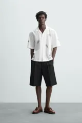 Camisa de manga corta confeccionada en mezcla de algodón y lino, color blanco. Presenta un cuello tipo bowling, calce regular y cierre frontal con botones. Destaca por sus bordados lineales en contraste de color negro con motivos de frutas y hojas distribuidos por toda la prenda.