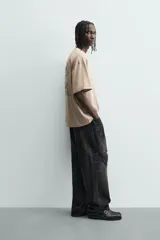 Camiseta de manga corta y cuello redondo, color beige claro, con corte relajado (relaxed fit) y un pequeño texto bordado en el pecho.