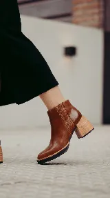 Bota corta de cuero color marrón con acabado liso, que presenta un detalle de textura acanalada en el lateral y cierre interno. Cuenta con un taco ancho y estable de color negro.