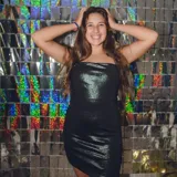 Vestido corto negro brillante, con escote recto, espalda descubierta con tiras cruzadas y falda ajustada con tajo lateral.