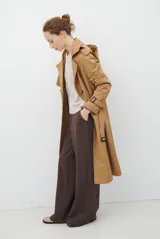 Trench coat color beige con cuello de solapa, cierre frontal abotonado, bolsillos laterales y mangas largas con cintas ajustables. Incluye capucha desmontable y forro interior rayado. La tela exterior cuenta con protección repelente a la lluvia y manchas.