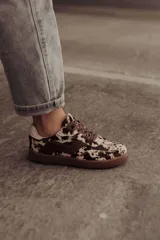 Zapatillas de cuero con estampado que simula pelo de vaca o manchas marrones y blancas. Tienen detalles en cuero marrón liso, cordones marrones y suela de goma color caramelo.