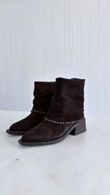 Bota de caña media confeccionada en cuero gamuzado color marrón oscuro, con diseño de caña arrugada y detalle de costura trenzada en la base. Cuenta con un taco cuadrado de madera y punta cuadrada.