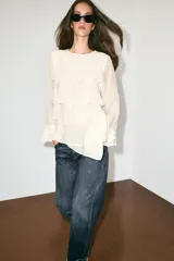 Blusa marrón de cuello redondo y manga larga, con volantes horizontales superpuestos y bajo asimétrico. Cuenta con detalle de abertura y botón en la espalda.