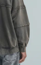 Buzo de cuello redondo, color gris oscuro con efecto desgastado (acid wash). Presenta costuras expuestas y detalles de roturas en los bordes. El calce es oversize y crop, con mangas largas abullonadas y puños y ruedo con terminación deshilachada y ondulada.