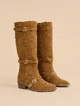Bota alta de gamuza color marrón, con caña ancha, taco bajo cuadrado y punta fina. Cuenta con tres tiras con hebillas metálicas doradas ajustables: una en la parte superior, otra a la altura del tobillo y otra sobre el empeine.
