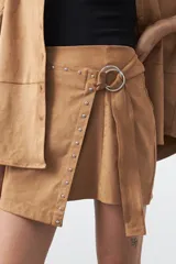 Minifalda de gamuza símil color marrón, con diseño envolvente tipo wrap, detalle de tachas metálicas en el frente y cinturón integrado con aro metálico.