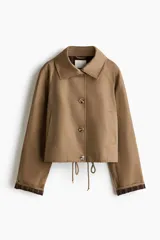 Chaqueta corta de sarga color marrón tostado, con cuello clásico, mangas raglán y cierre frontal con botones. Cuenta con bolsillos laterales, cordón ajustable en el bajo y puños con vuelta que dejan ver un forro a cuadros.