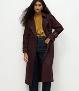 Trench coat largo de gamuza sintética con cuello peaked, cierre frontal cruzado con botones y cinturón ajustable en la cintura. Presenta mangas largas con reguladores en los puños y forro interior.