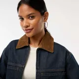 Chaqueta vaquera de color azul oscuro, corte corto y holgado, con cierre frontal de cremallera y dos bolsillos grandes con solapa en el frente. Posee cuello de pana marrón.