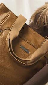 Cartera tipo tote marrón de diseño práctico y versátil, confeccionada en símil cuero con textura granulada. Cuenta con asas cortas fijas, bolsillos exteriores con cierre al frente y atrás, y bolsillos laterales abiertos. Su interior es amplio e incluye bolsillos organizadores. Viene con una correa ajustable y desmontable.
