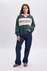 Sudadera polo verde oscuro con cuello y franja central en color beige. Presenta un diseño de manga larga y cuenta con un bordado frontal con el texto "BROOKLYN ATHLETICS DEPT. Est. 1995".