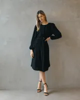 Vestido midi negro de silueta fluida, con mangas largas abullonadas y cintura marcada mediante un cinturón incorporado con hebilla metálica. Presenta un diseño de escote cerrado y caída liviana.