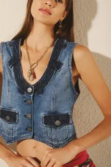 Chaleco de denim rígido azul con efecto desgastado, corte ajustado y escote en V. Presenta botones metálicos grabados en el frente, dos bolsillos con tapa en el pecho y un detalle de tira de ajuste en la espalda.