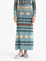 Pollera larga de punto con diseño a cuadros en tonos verde, marrón, negro y beige. Presenta un corte ajustado en la cadera que se ensancha hacia el ruedo y una cintura con detalle de hilo metálico.