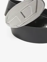 Cinturón de cuero negro con hebilla grande y llamativa de metal plateado con el logo de Diesel en forma de 'D' estilizada.
