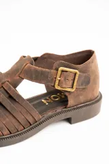 Sandalias cangrejeras de cuero color marrón, con diseño de tiras cruzadas y cierre de hebilla metálica en el lateral. Poseen suela plana y gruesa.
