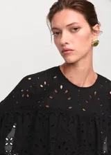 Blusa negra de manga larga con diseño de volantes y bordados perforados. Presenta cuello redondo con cierre de lazo y corte recto.