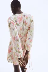 Vestido corto con textura plisada, estampado floral en tonos pastel (rosa, verde, amarillo, azul) sobre fondo crema. Presenta escote pronunciado en V con fruncido central y mangas globo largas ajustadas con lazos en los puños.