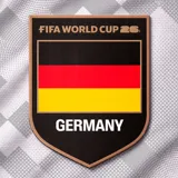Camiseta de fútbol blanca con diseño geométrico sutil, cuello redondo negro y detalles en naranja. Presenta el logo de la FIFA World Cup 26 y el escudo de Alemania en el pecho.