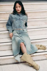 Falda midi de denim con lavado claro, tiro medio y cintura cruzada con cierre asimétrico de cremallera y botones metálicos. Presenta bolsillos delanteros y bajo asimétrico.