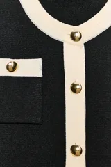 Chaqueta de punto de corte recto y manga larga, con cuello redondo. Presenta un diseño bicolor en negro y crema, con cierre frontal de botones dorados y bolsillos de plastrón en el delantero con detalles en contraste.