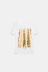 Camiseta de algodón con cuello redondo y manga corta. Presenta un diseño de estampación metalizada en color dorado en la parte frontal con detalles de texto.