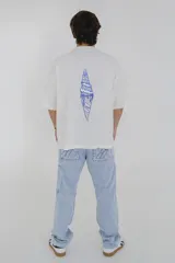 Remera color crema de corte holgado con cuello redondo y mangas cortas. Estampado en el pecho con la inscripción "Pulau Cold Winter" en letras azules y amarillas.