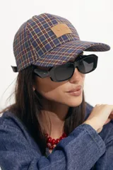 Gorra tipo baseball con estampado de cuadros escoceses en tonos azul, naranja y beige. Presenta un parche de cuero sintético en el frente y cierre ajustable en la parte posterior.