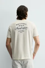 Camiseta de corte relajado, color crudo, con cuello redondo y manga corta. Presenta un pequeño estampado gráfico en el pecho con la inscripción 'Whitsunday Island'.