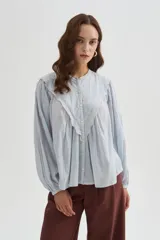 Blusa de algodón color beige, con cuello redondo y cierre frontal con botones. Presenta delicados bordados decorativos en tono tostado sobre el pecho y los hombros, mangas largas abullonadas con puños ajustados y diseño holgado.