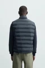 Chaleco acolchado verde oscuro, con cuello alto, cierre frontal con cremallera y bolsillos laterales. Confeccionado en tejido técnico repelente al agua.