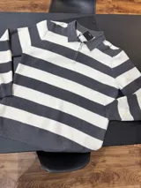 Buzo tejido de punto a rayas horizontales en color gris y blanco, con cuello polo y medio cierre frontal. Presenta puños y ruedo acanalados.