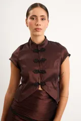 Top de tafeta elastizada con inspiración oriental, color marrón, cuello mao y cierre frontal con nudos chinos y broches. Presenta mangas cortas con detalles de puntilla negra en los bordes.