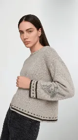 Sweater de lana color beige con calce holgado y cuello redondo. Presenta un bordado negro con diseño de arabescos en una de las mangas y detalles de rayas y puntos en contraste en el ruedo y los puños.