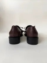 Mocasín de cuero marrón con textura granulada, diseño de horma cuadrada, detalle de flecos y lazo decorativo en el empeine, montado sobre una suela y taco bajo de color negro.