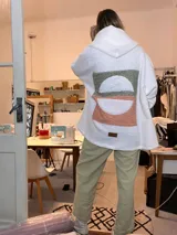 Campera de corderito blanco con capucha, de corte holgado y diseño largo. Presenta un aplique rectangular en la espalda con diseño geométrico de semicírculos en tonos verde y naranja con estampado floral pequeño.