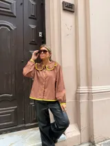 Camisa de color marrón chocolate con cuello redondo grande y ribetes en color amarillo vibrante. Presenta cierre frontal con botones amarillos, mangas largas con puños elásticos y un corte holgado.