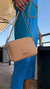 Bandolera pequeña de diseño rectangular en color beige, confeccionada en cuero sintético. Presenta una correa larga ajustable del mismo material y cierre superior con cremallera. Incluye el logo de la marca grabado en la parte frontal.