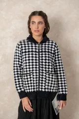 Chaqueta de punto grueso con patrón geométrico de cuadros en blanco y negro. Presenta cuello camisero, cierre frontal con cremallera y tirador circular, bolsillos frontales y puños acanalados.