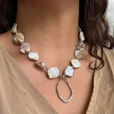 Collar corto con cuentas irregulares de nácar en tonos blancos y beige, separadas por pequeñas cuentas metálicas plateadas. Incluye un dije colgante de metal plateado con forma orgánica irregular.