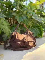 Bolso de cuero marrón con diseño tipo morral, cuenta con doble correa de hombro y correa larga ajustable, cierre superior y detalles de remaches metálicos.