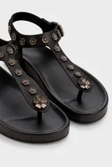 Sandalia baja de cuero vacuno color negro, tipo thong sandal, con tira vertical en el empeine decorada con apliques metálicos en forma de flores y tira ajustable en el talón con hebilla.