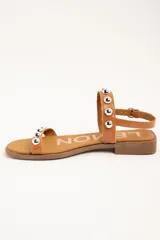Sandalias planas de cuero color suela, con tira en la puntera y tira en el empeine decoradas con tachas semiesféricas plateadas, y tira ajustable con hebilla en el tobillo.