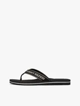 Ojotas negras con tiras de textil trenzado que presentan el logo de Tommy Hilfiger en jacquard con detalles en blanco y dorado. La suela es de EVA dentada con una entresuela a contraste blanca.