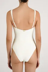 Traje de baño enterizo color crema con diseño bandeau, fruncidos laterales y tres aberturas circulares frontales decoradas con detalles metálicos brillantes.